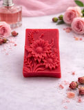 Red floral soap bar on a light surface with pink roses and a perfume bottle in the background.
