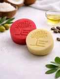 Two natural soap bars, one red and one yellow, on a light surface with olive branches and bowls of ingredients.