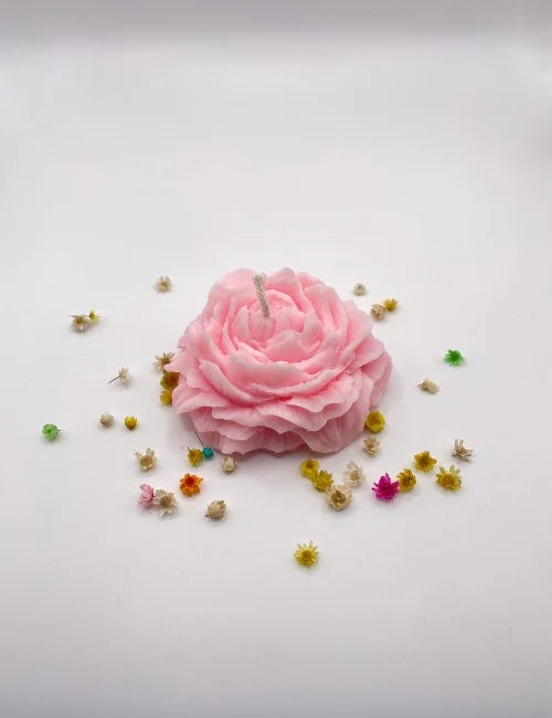 A light pink natural candle in a rose shape, surrounded by small decorative flowers.